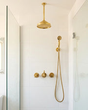 Unlacquered brass ceiling mount shower system with round rain shower head and handheld shower in a modern tiled bathroom.