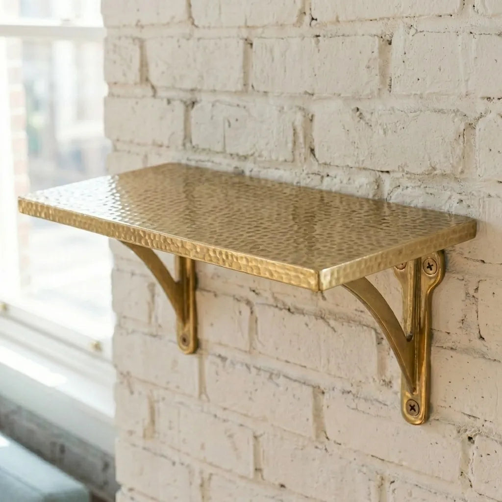 Handmade hammered brass wall shelf mounted on a painted brick wall, featuring a textured metal surface and decorative support brackets