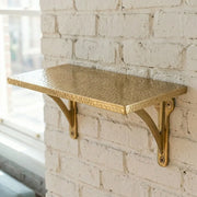 Handmade hammered brass wall shelf mounted on a painted brick wall, featuring a textured metal surface and decorative support brackets