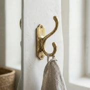Traditional brass double wall hook with decorative backplate mounted on a white wall holding a linen towel.