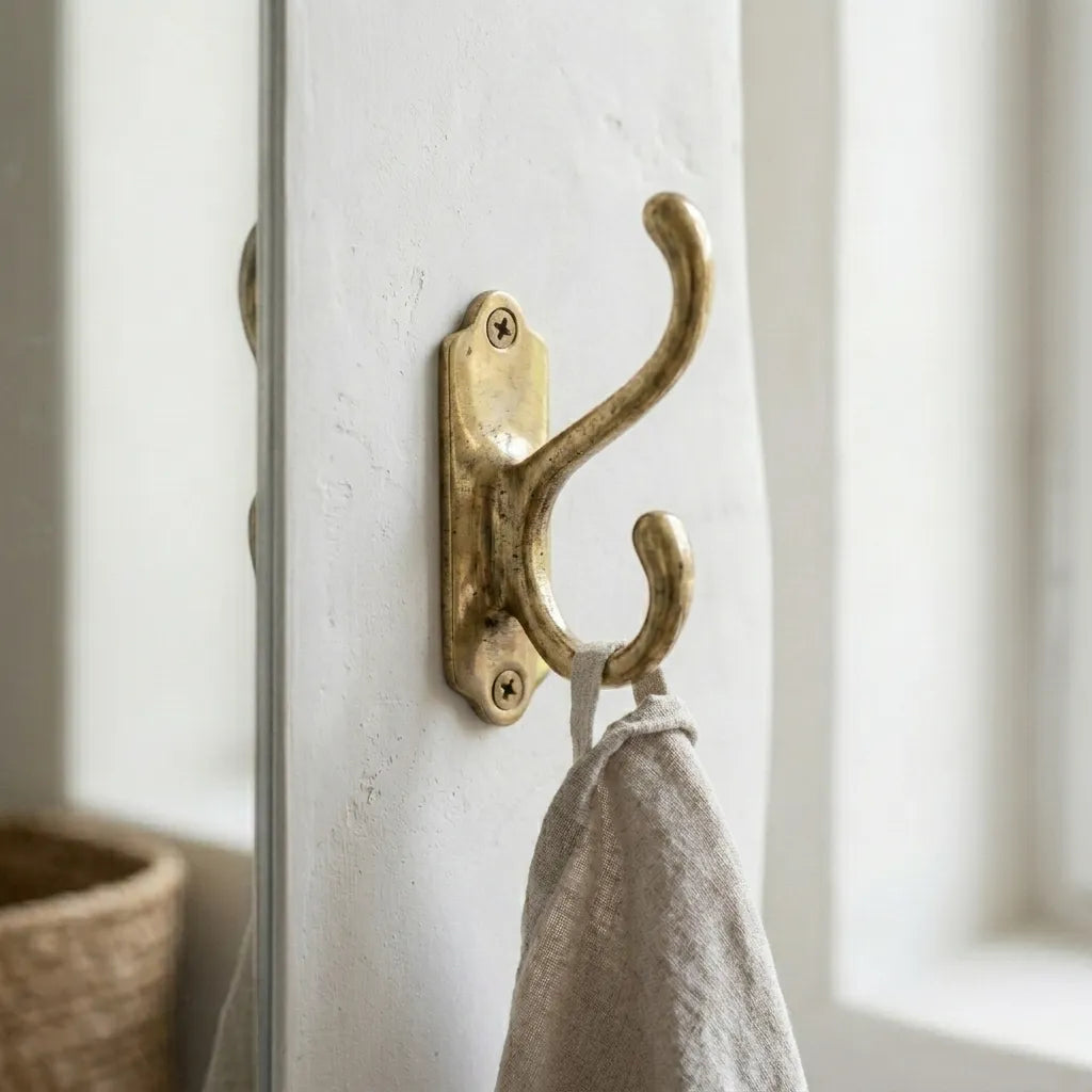 Traditional brass double wall hook with decorative backplate mounted on a white wall holding a linen towel.