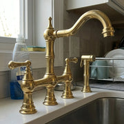A polished brass bridge faucet set installed on a white kitchen sink countertop, with a matching side sprayer and a soap dispenser nearby in a real kitchen setting.