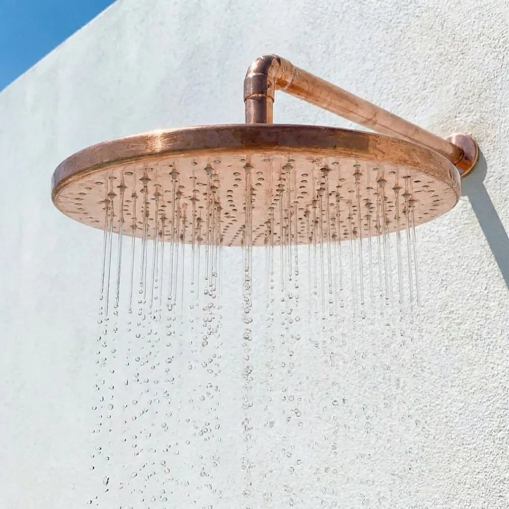 Handcrafted round copper rainfall shower head with exposed wall arm installed in a modern outdoor shower.