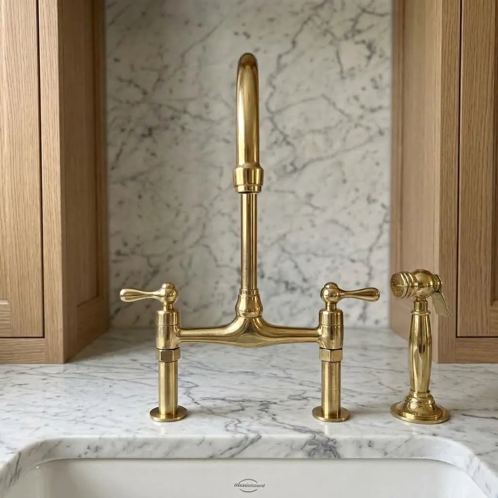 Unlacquered brass bridge faucet with a tall gooseneck spout and two separate handles, installed on a white marble countertop with a matching marble backsplash and light wood cabinets.