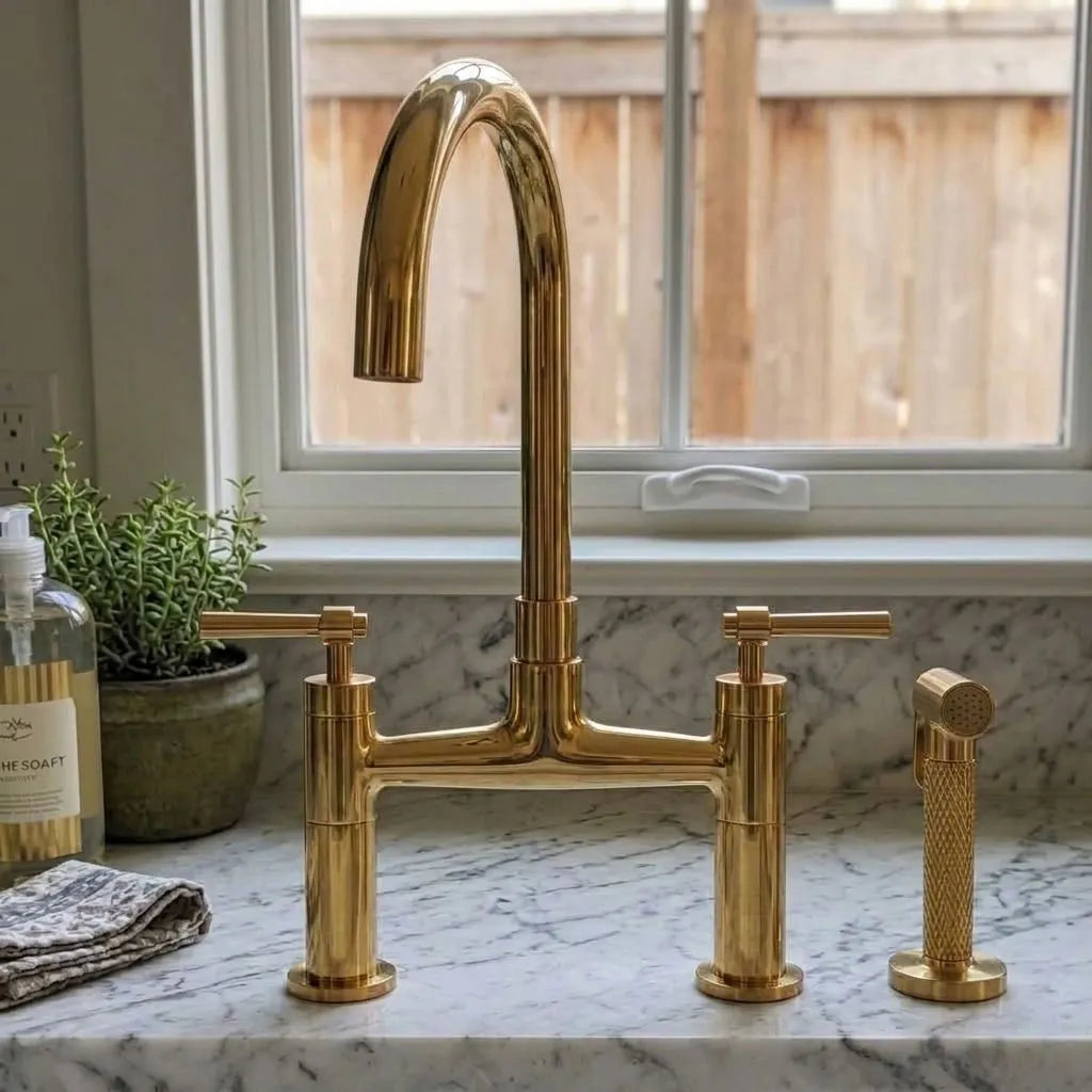 A polished brass bridge faucet set with a high-arc spout, two cross-handle valves, and a matching handheld sprayer, installed on a white marble kitchen countertop in front of a window.