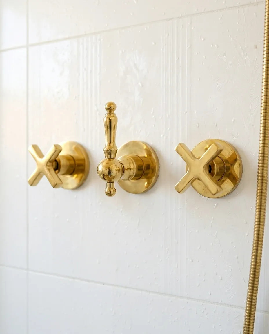 Unlacquered brass concealed shower valve with cross handles and central diverter installed on a white tiled shower wall.