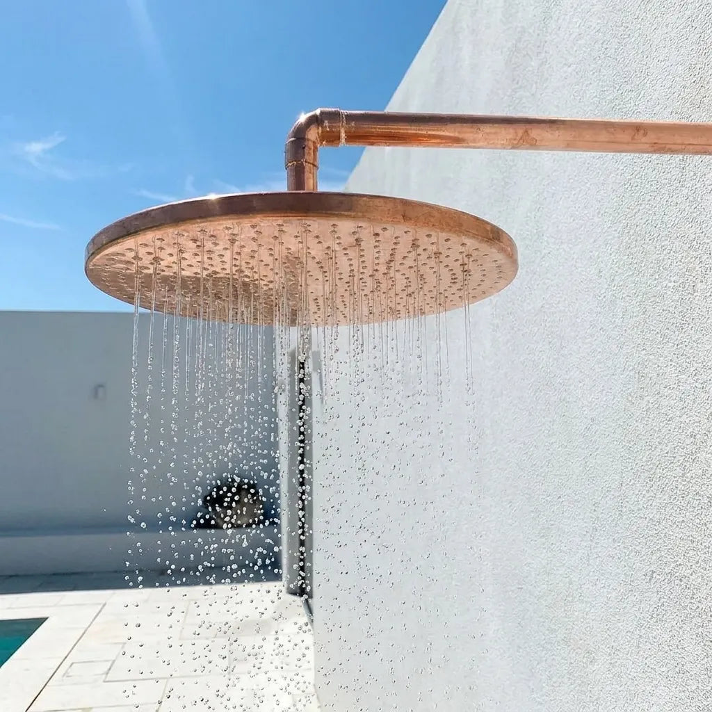 Large hammered copper rain shower head with wall-mounted arm delivering a luxurious rainfall shower experience outdoors.