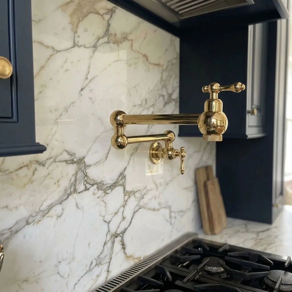 A polished brass wall-mounted kitchen faucet with an adjustable arm and cross-handle, installed on a white marble backsplash in a modern kitchen with dark blue cabinets.