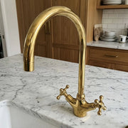 A polished brass kitchen faucet with a high-arc spout and cross-handle valves, installed on a white marble countertop in a kitchen setting.