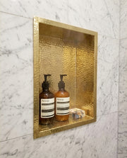 Hand hammered solid brass recessed shower niche installed in a marble tiled bathroom wall.