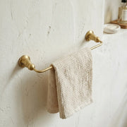 Unlacquered brass hand towel bar mounted on a textured bathroom wall with a soft woven towel draped across the rail