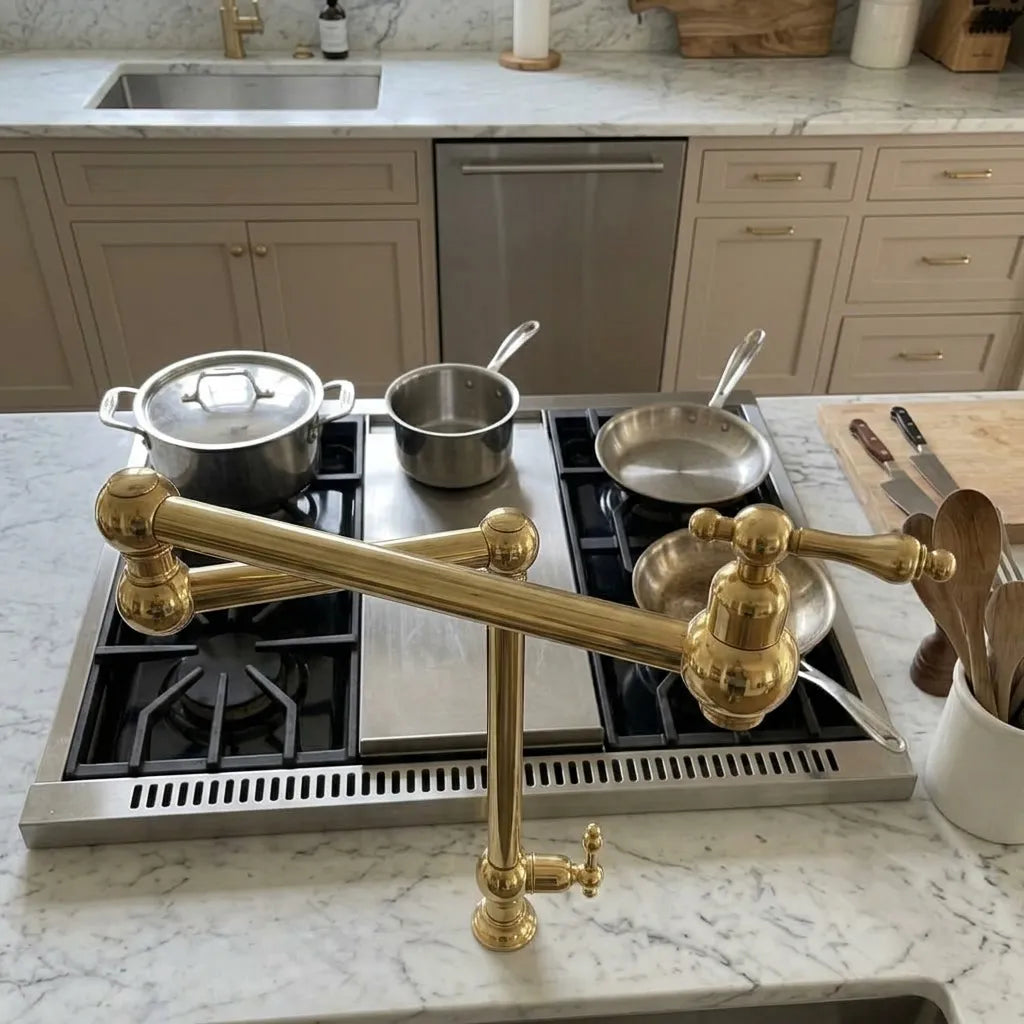 A polished brass kitchen faucet with an adjustable gooseneck spout is mounted on a white marble countertop in a modern kitchen setting with a gas stove, pots, and utensils.