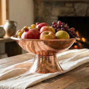 Handcrafted hammered copper pedestal fruit bowl filled with fresh fruit on a rustic farmhouse dining table.