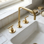A three-hole bathroom faucet set in brushed brass with a high-arc spout and two lever handles, installed on a white stone sink with tiled walls.