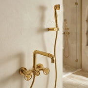 Gold-finished wall-mounted bathroom faucet and handheld shower set installed on a textured off-white wall, with a glass shower door visible in the background.
