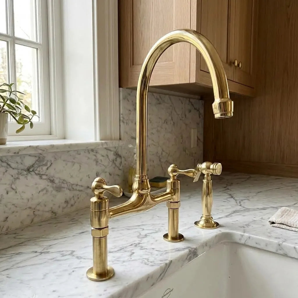 Unlacquered brass bridge faucet with a high-arc spout and two separate lever handles, installed on a white marble kitchen sink countertop.