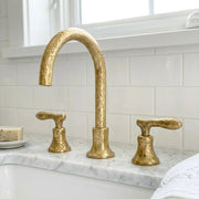 Hammered brass widespread bathroom faucet with textured arch spout and sculpted lever handles on a marble vanity