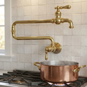 A polished brass wall-mounted kitchen faucet with a curved gooseneck spout and separate handle, shown above a copper pot on a gas stovetop against a white tiled backsplash.