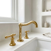 Handcrafted brass bridge faucet with sculpted curved spout and dual lever handles installed on a white countertop basin