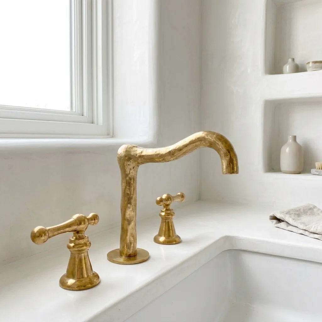 Handcrafted brass bridge faucet with sculpted curved spout and dual lever handles installed on a white countertop basin