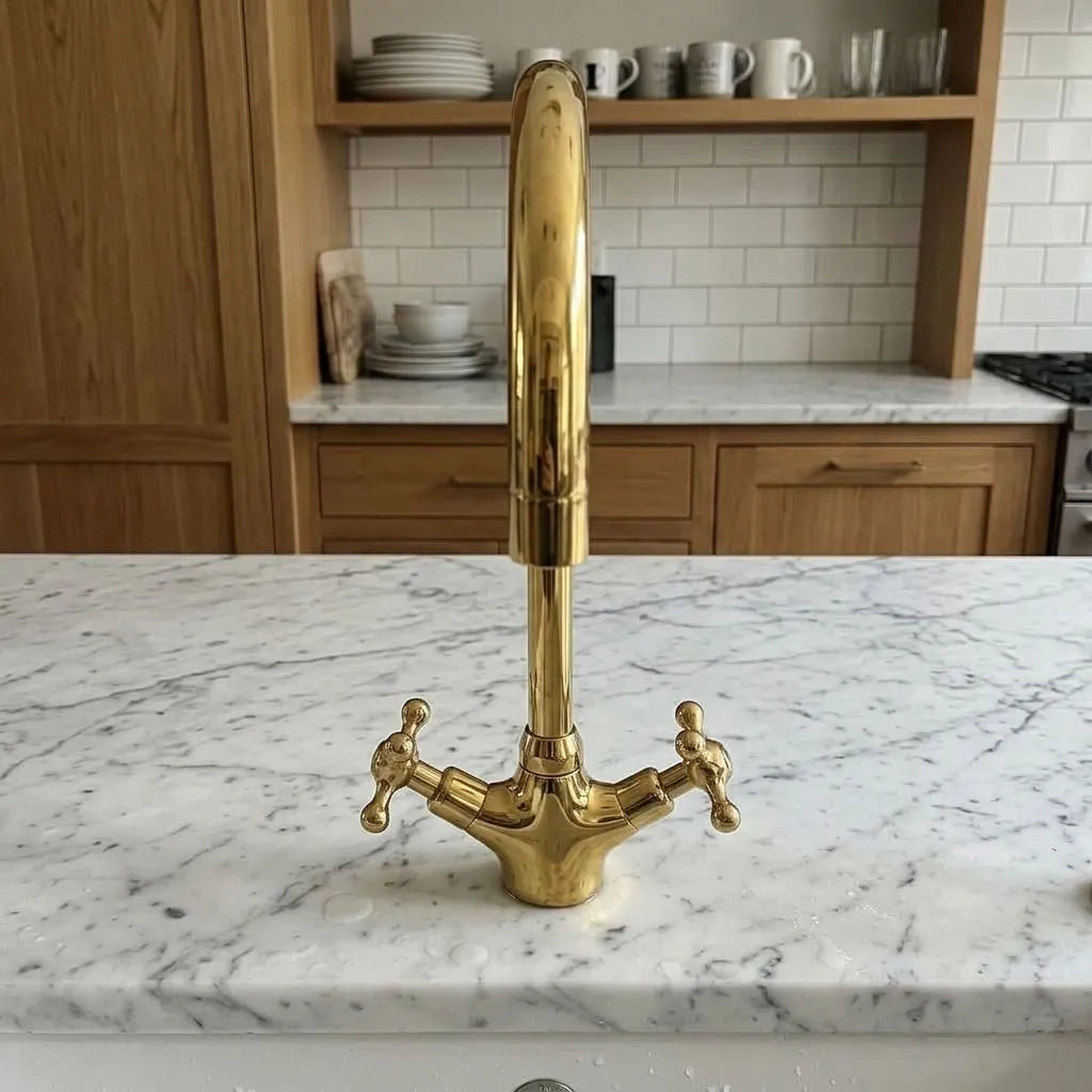 A polished brass kitchen faucet with a tall curved spout and two cross handles, set on a white marble countertop in a modern kitchen with wood cabinets and subway tiles.