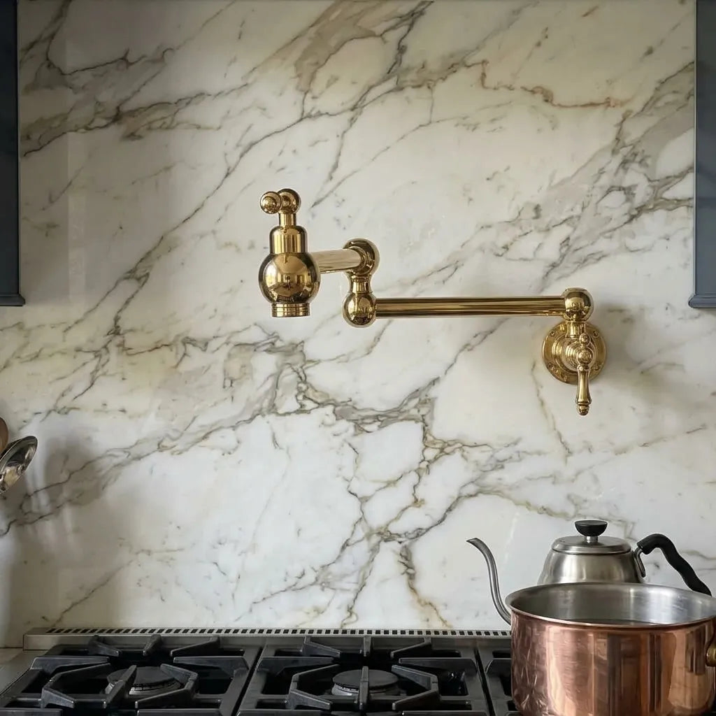 A polished brass wall-mounted kitchen faucet with a curved arm and separate spout, installed on a white marble backsplash above a gas stove with a copper pot and stainless steel kettle.