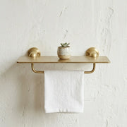 Solid brass bathroom shelf with integrated towel rail mounted on a textured wall, featuring a minimalist storage ledge and hanging towel
