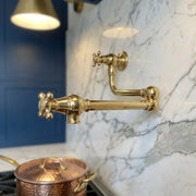 A polished brass wall-mounted kitchen faucet with a cross-handle valve and flexible gooseneck spout, installed on a white marble backsplash next to a blue cabinet and a copper pot on a stove.