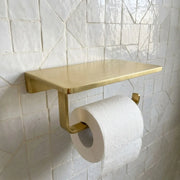 A modern wall-mounted toilet paper holder with a rectangular shelf and roll mount in a brushed gold finish, shown holding a white toilet paper roll against a white tiled wall.