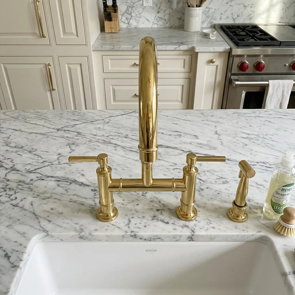 A polished gold bridge faucet set with a tall central spout and two separate handles, installed on a white and gray marble kitchen sink countertop in a modern kitchen.