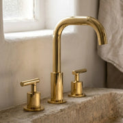 A polished brass three-hole bathroom faucet set with a high-arc spout and two separate lever handles, installed on a stone sink next to a window.