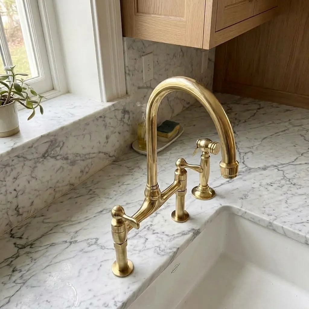 Unlacquered brass bridge faucet with a high-arc spout and two lever handles, installed on a white marble kitchen sink countertop.