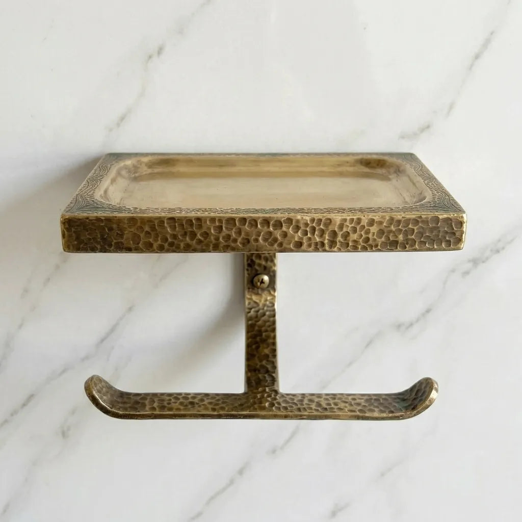Wall mounted brass toilet roll holder featuring a textured hammered finish and upper tray for toiletries, candles, or decorative accents