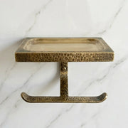 Wall mounted brass toilet roll holder featuring a textured hammered finish and upper tray for toiletries, candles, or decorative accents