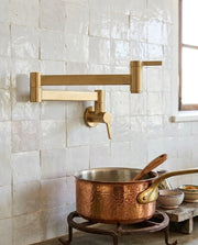 A brass wall-mounted kitchen faucet with an adjustable arm and single lever handle, installed on a white tiled wall above a copper pot on a burner.