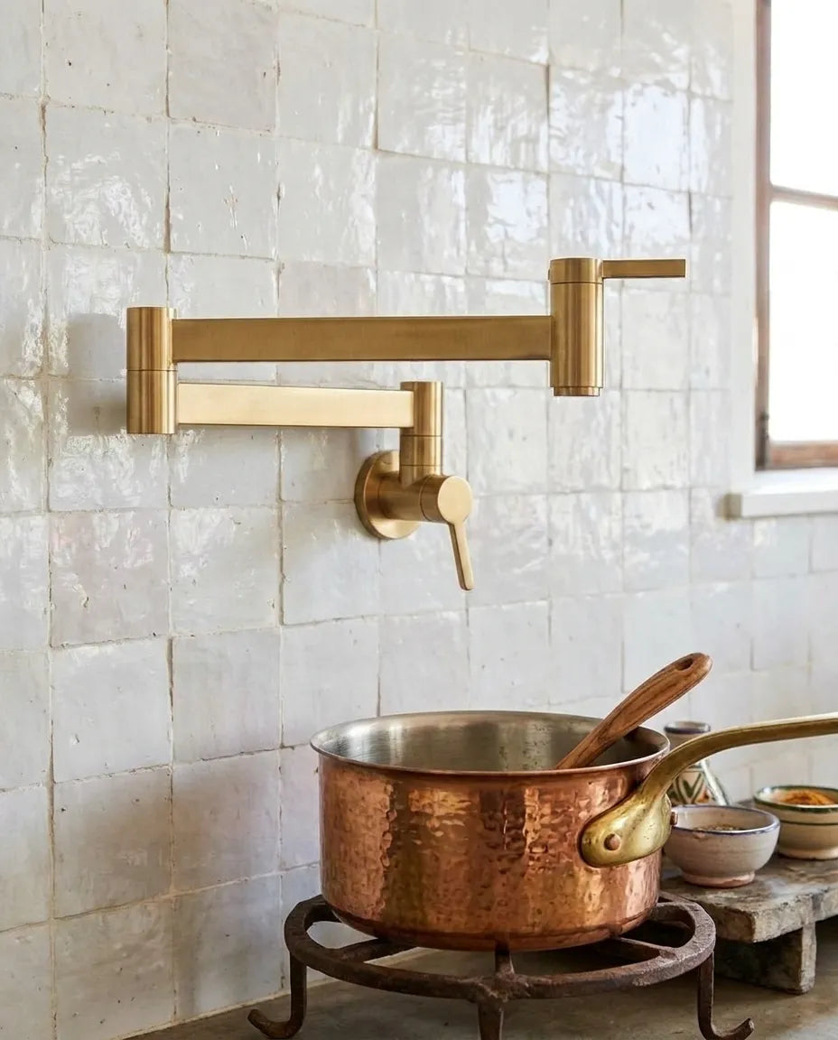 A brass wall-mounted kitchen faucet with an adjustable arm and single lever handle, installed on a white tiled wall above a copper pot on a burner.