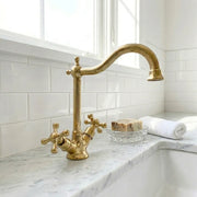 Unlacquered brass bridge kitchen faucet with high arc swan neck spout and classic cross handles, installed on a marble countertop
