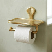 Solid brass toilet paper holder with storage shelf mounted on bathroom wall in a warm unlacquered brass finish