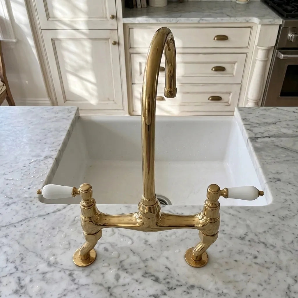 Unlacquered brass bridge faucet with two white ceramic handles installed in a white undermount sink on a white and gray marble kitchen countertop, with white cabinets in the background.