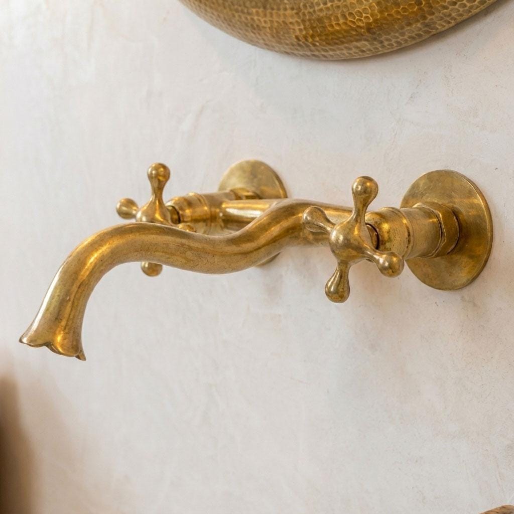 Unlacquered brass wall mount bathroom faucet with curved spout and traditional cross handles.