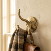 Hand forged brass triple wall hook mounted on a textured wall holding a scarf and leather bag.