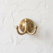 Unlacquered brass double wall hook mounted on a textured wall.